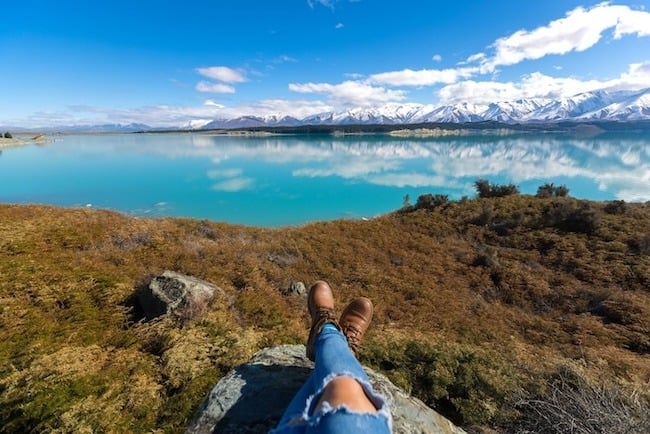 A person's legs while sitting on rocks with Lake Pukaki in the background